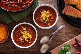Bowls of chili with cornbread, cilantro, peppers, and cheese on the side