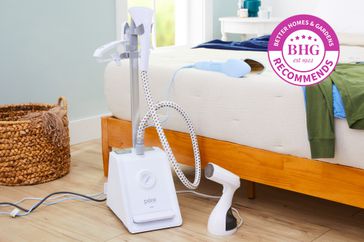 The Pure Enrichment PureSteam Pro Upright Clothes Steamer on a wood floor next to a bed with clothes laying on it.